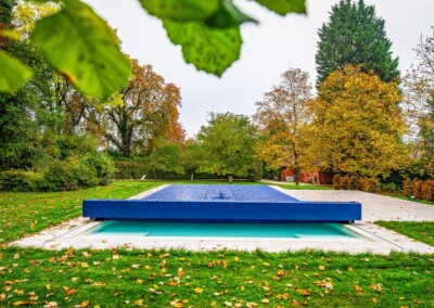 BLUE-COVER-(17)-min Ein blauer Pool mit Abdeckung in einem herbstlichen Garten, umgeben von Bäumen und Laub auf dem Rasen.