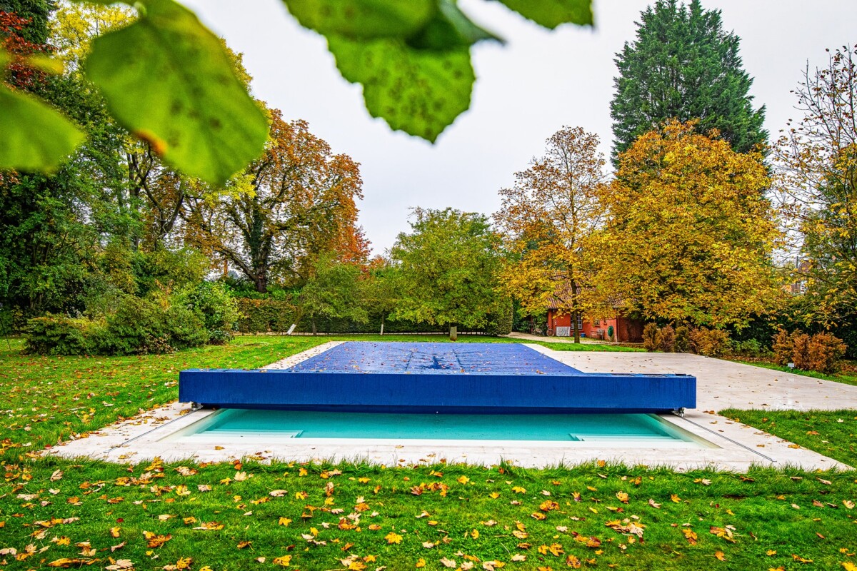 Ein blauer Pool mit Abdeckung in einem herbstlichen Garten, umgeben von Bäumen und Laub auf dem Rasen.