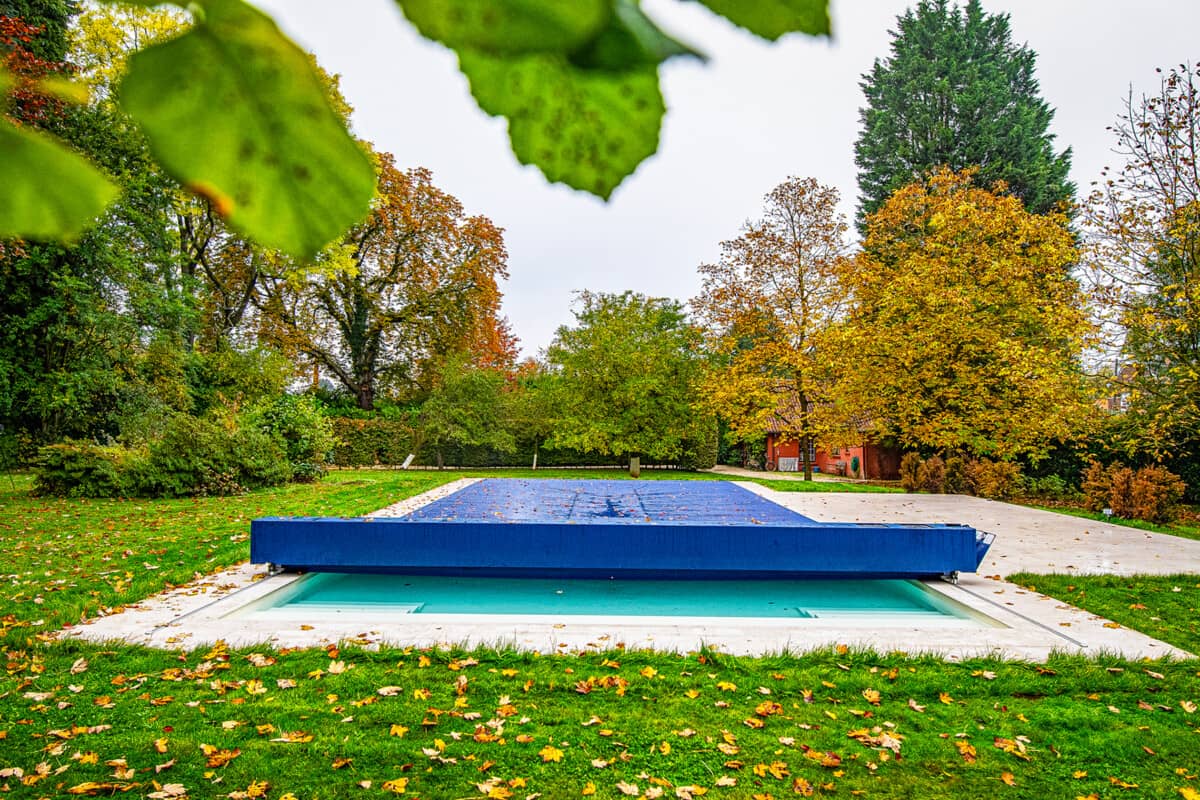Une piscine recouverte d'une bâche bleue est entourée d'arbres aux feuilles automnales dans un jardin verdoyant.
