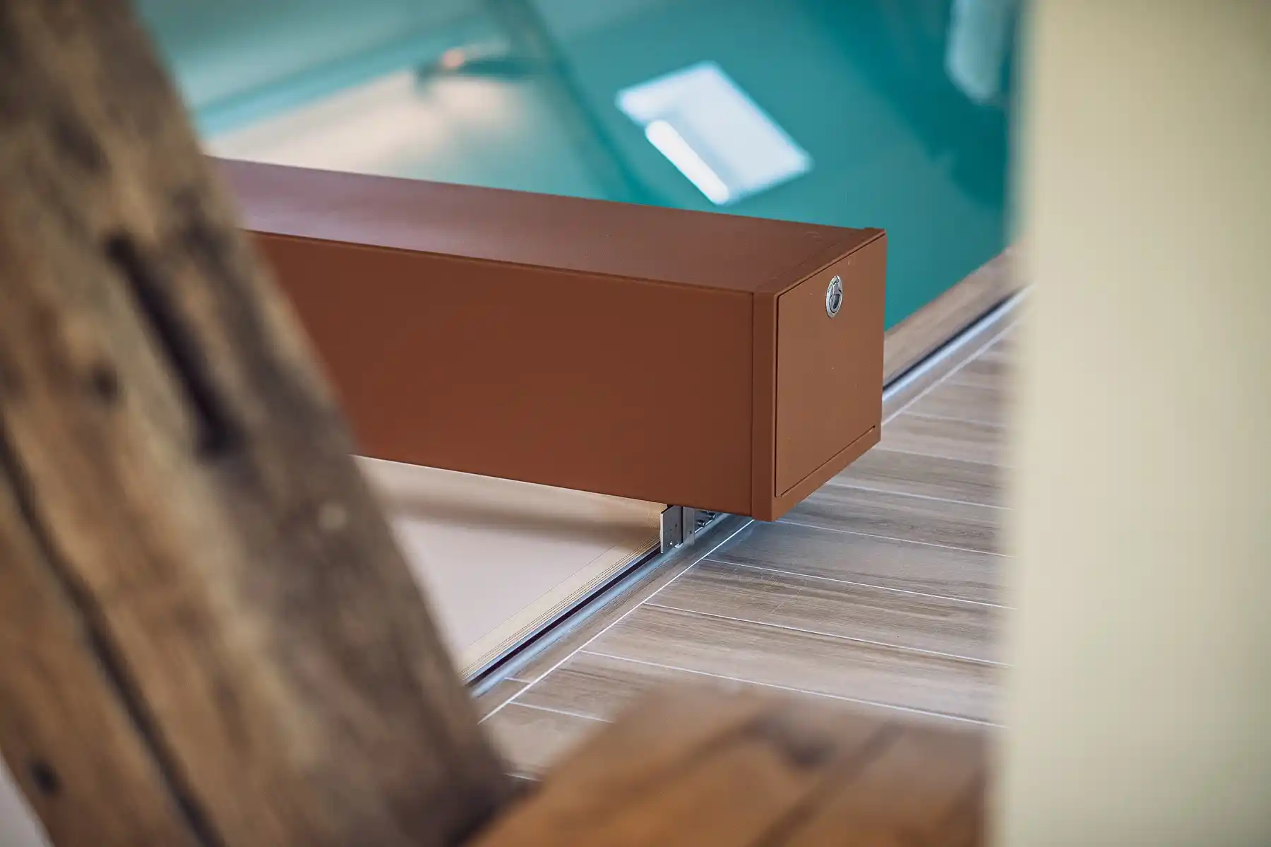 chez-alexandra-and-damien-2 Boîtier rectangulaire marron fixé sur un rail en métal, posé sur un sol en bois clair, avec une piscine en arrière-plan.