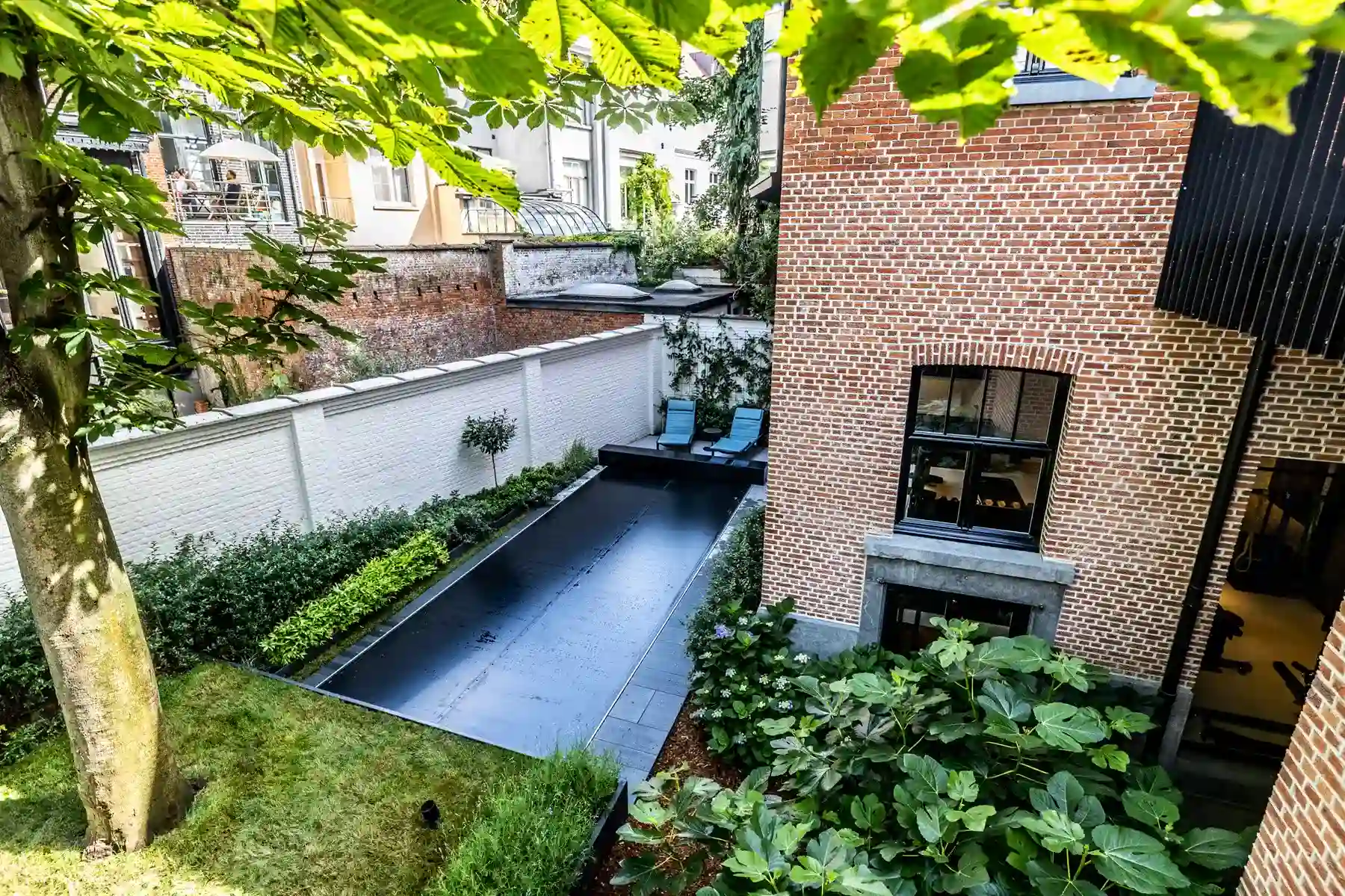 Chez-Anna-et-Luc-3 Cour arrière avec piscine couverte noire, entourée de murs en briques rouges et de végétation, deux chaises longues bleues visibles.