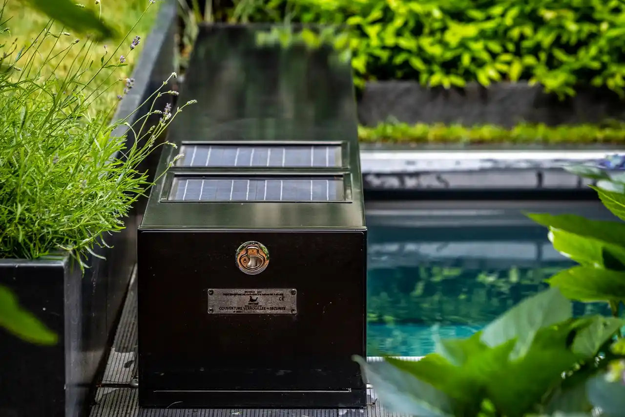 Chez-Anna-et-Luc-4 Boîtier noir avec panneaux solaires à côté d'une piscine, entouré de plantes vertes et de fleurs violettes.