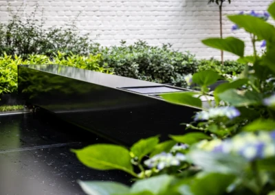 Chez-Anna-et-Luc-6 Une couverture de piscine noire entourée de plantes vertes et de fleurs violettes, avec un mur en briques blanches en arrière-plan.