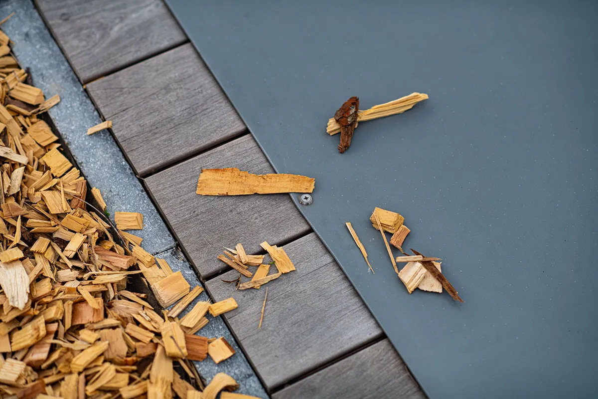 comment-faire-pour-mettre-sa-piscine-en-hivernage Des éclats de bois sont dispersés sur une couverture de piscine grise, à côté de dalles en bois.