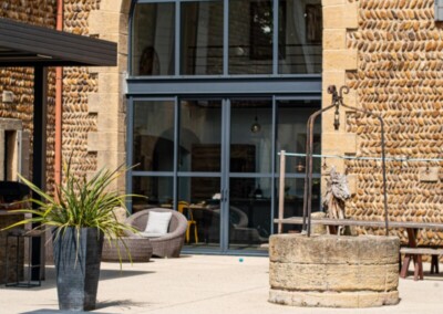 Travelhouse_10-min Façade en pierre avec grande fenêtre arquée, terrasse avec mobilier de jardin, plante en pot et piscine couverte par une structure grise.