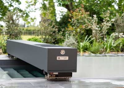 Couverture de piscine grise automatisée sur une terrasse en pierre, entourée de végétation verdoyante.