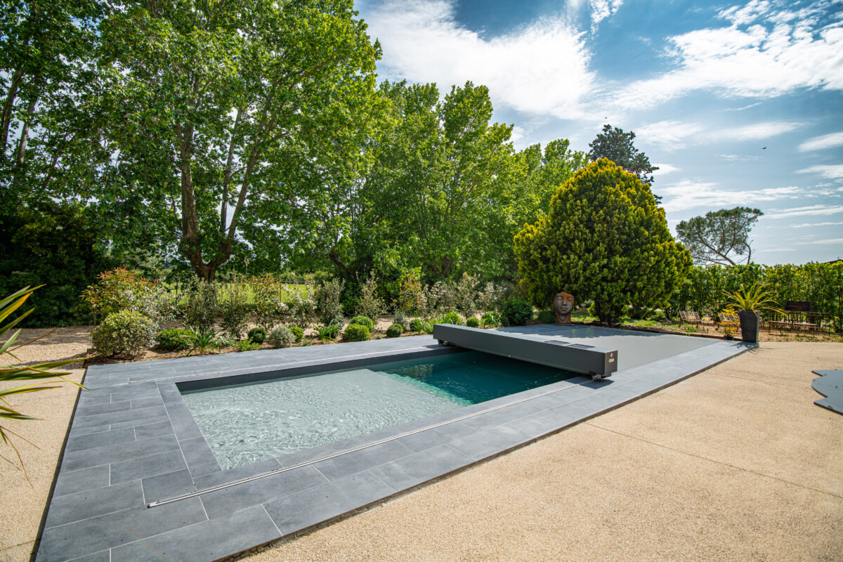 Travelhouse_27- Site Réal Piscine rectangulaire entourée de dalles grises, avec un volet de protection, bordée de végétation luxuriante sous un ciel partiellement nuageux.