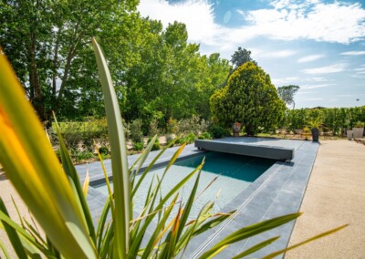 Travelhouse_28-min Piscine extérieure avec couverture grise entourée de végétation luxuriante et terrasse en pierre sous un ciel partiellement nuageux.