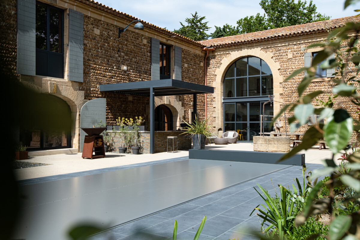 Travelhouse_37- Réal. Stéphane & Stéfany Cour intérieure avec façade en pierre, pergola grise, mobilier de jardin et couverture de piscine grise entourée de plantes vertes.