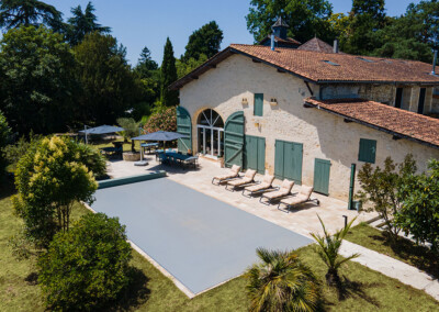 Sequoia1 Piscine rectangulaire couverte par une Coverseal grise, entourée de transats et d’une maison en pierre au style traditionnel avec volets verts.