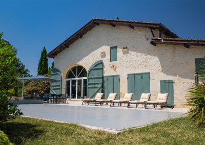 Sequoia2 Perspective rapprochée sur la piscine recouverte par une Coverseal grise, devant une maison de campagne avec volets verts et terrasse ensoleillée.